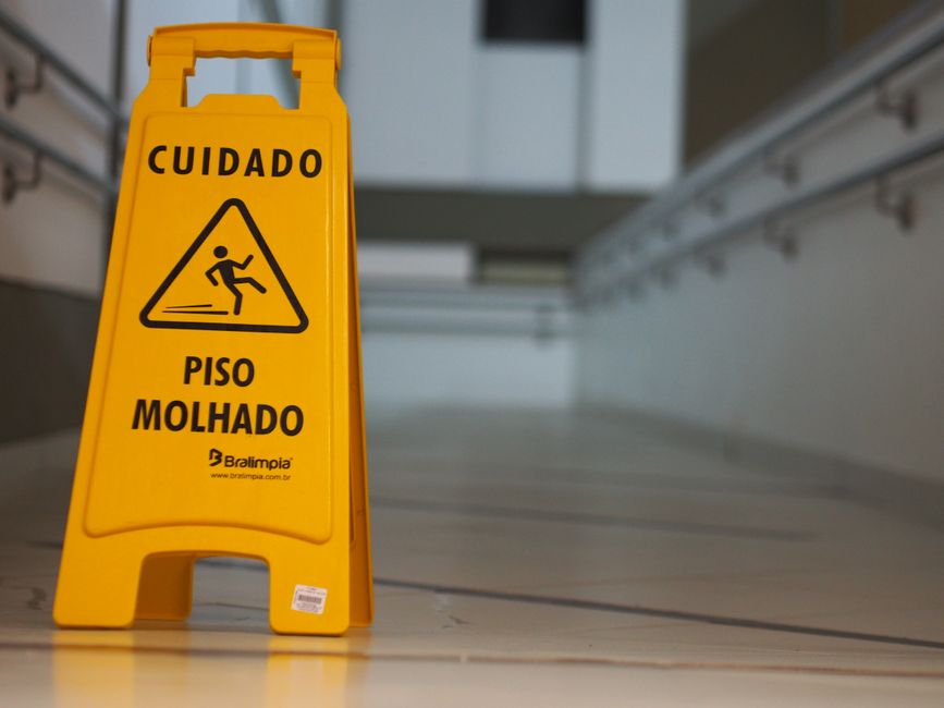 A yellow wet floor warning sign with the word 'Cuidado' at the top and an icon of a person slipping, positioned on a light-colored tiled floor in a commercial cleaning setting. The sign is placed near