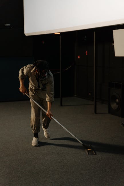 A person dressed in beige work overalls and white shoes is engaged in surface cleaning on a carpeted floor within an indoor space. They are using a long-handled tool to vacuum or sweep the carpet, whi