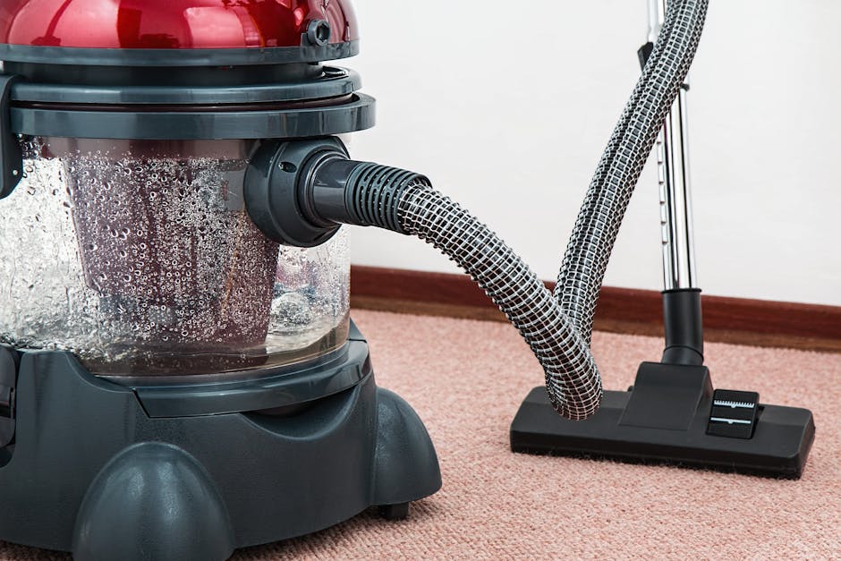 A person dressed in beige work overalls and white shoes is engaged in surface cleaning on a carpeted floor within an indoor space. They are using a long-handled tool to vacuum or sweep the carpet, whi