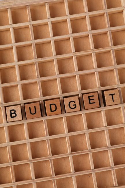 A wooden compartmentalized tray with multiple small square sections, predominantly in light natural wood tone. In the lower part of the tray, there are six small wooden blocks spelling the word 'BUDGE